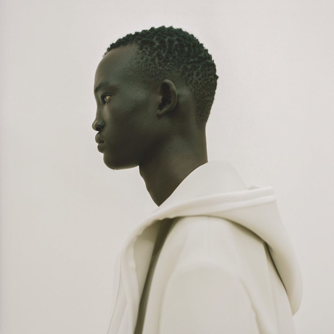 Profile of an adult male of African descent wearing a white hooded sweatshirt in a softly lit indoor studio environment.