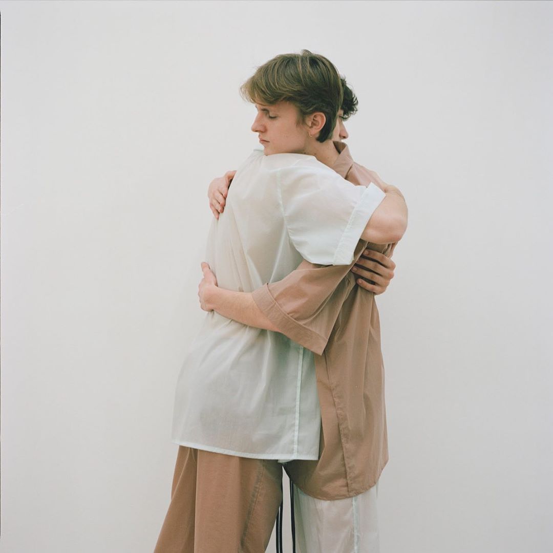 Two young adults embracing indoors against a white wall, wearing casual brown and off-white outfits with soft, diffused lighting.