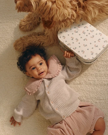Overhead shot of a baby lying on a rug next to a tan dog and a small floral suitcase. The baby wears a pink cardigan and shorts.