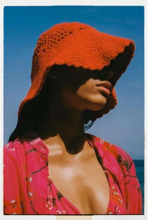 Model wearing a textured orange crochet bucket hat and pink floral shirt outdoors against a bright blue sky.
