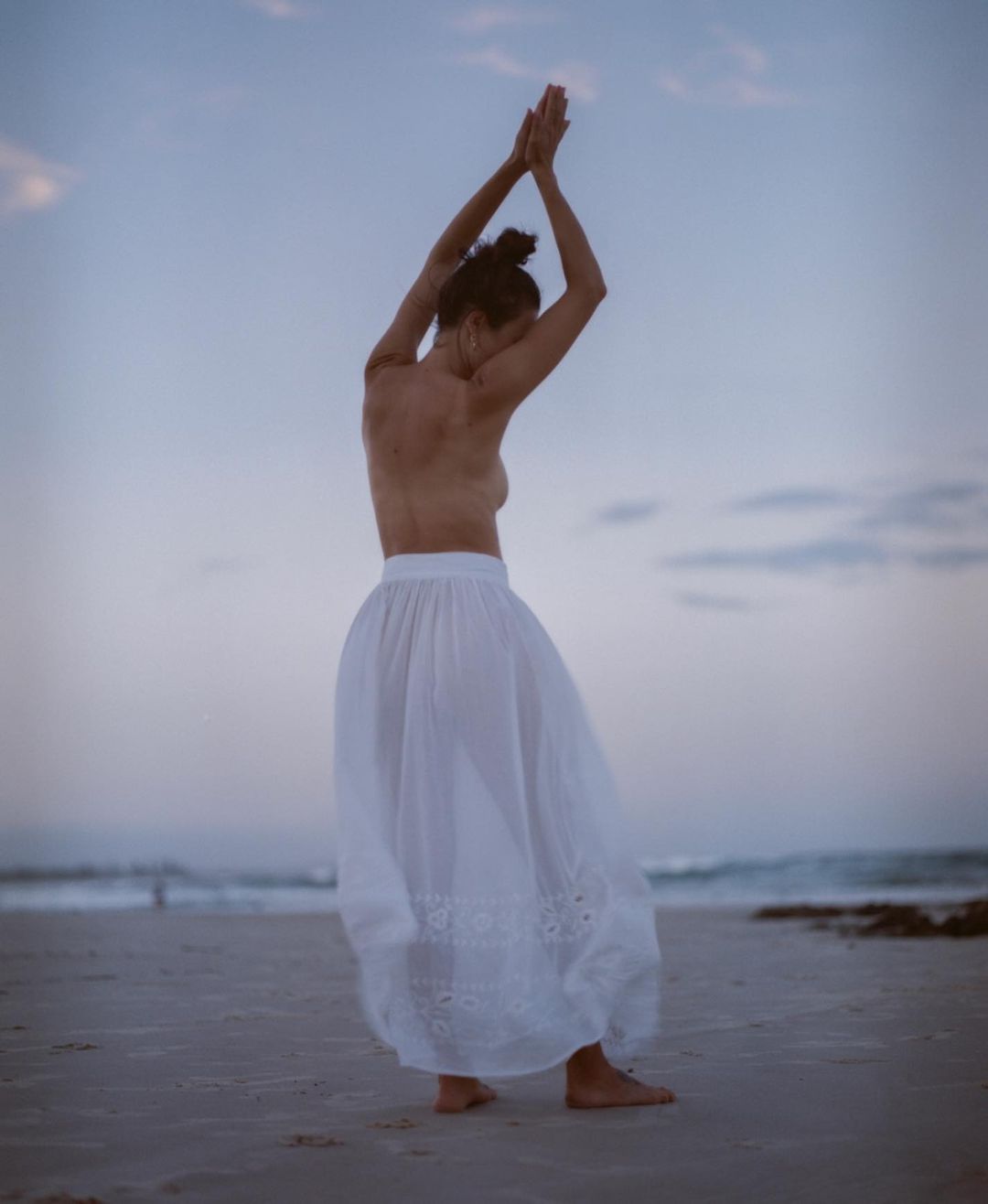 Woman in a long white skirt poses on a beach at dusk with arms raised in a gesture of freedom and dance as ocean waves roll in.