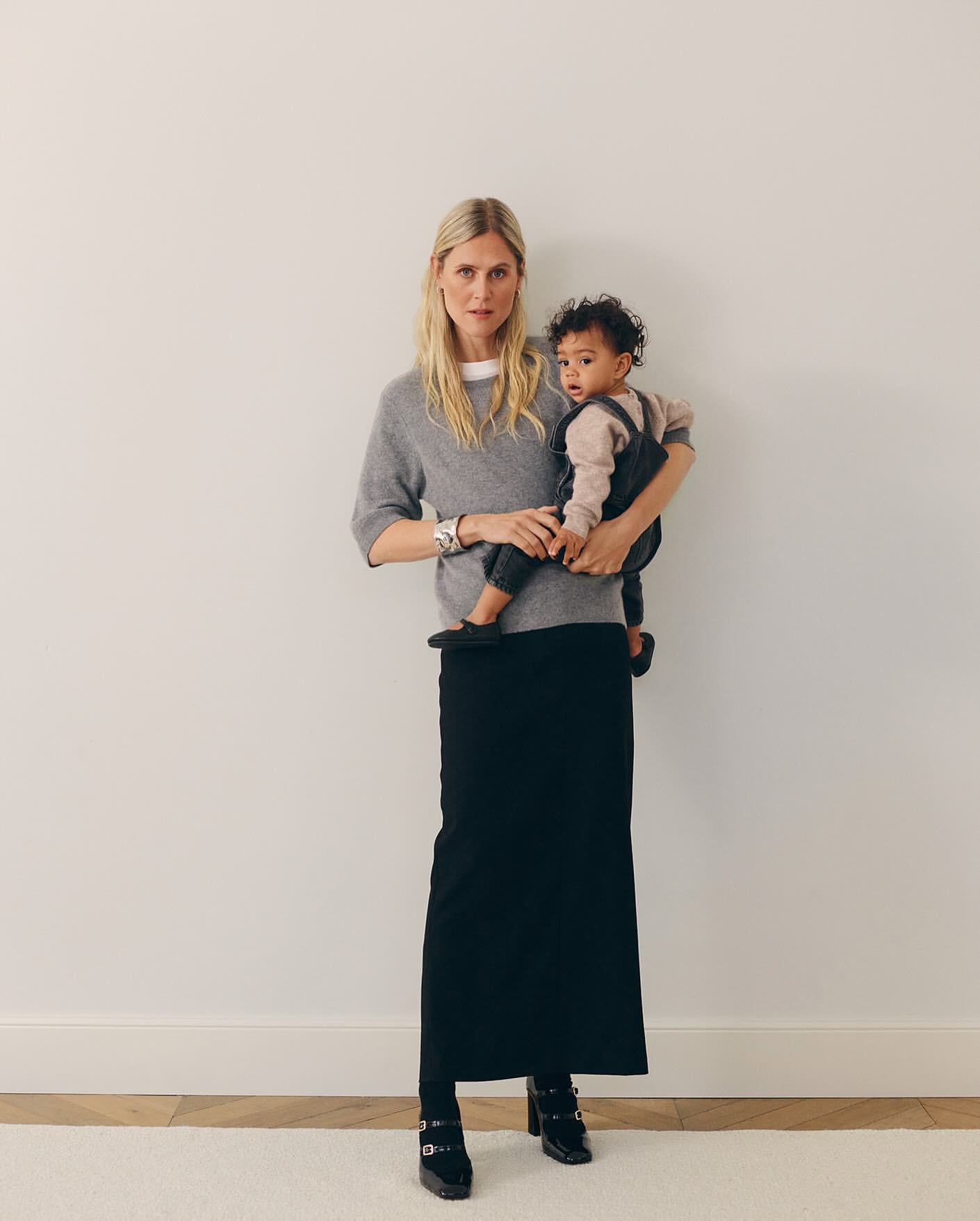 A mother wearing a gray sweater and black skirt holds her infant, who is wearing a brown shirt and black overalls, in an indoor space.