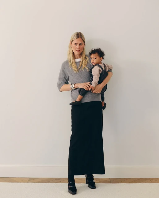 A mother wearing a gray sweater and black skirt holds her infant, who is wearing a brown shirt and black overalls, in an indoor space.