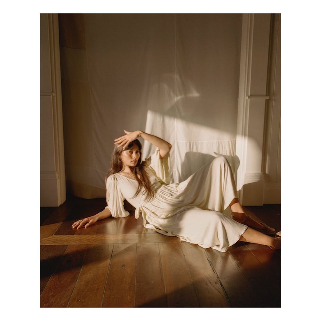 Adult woman in dress lying on wooden floor in indoor setting, casting shadow on face, bathed in natural sunlight near white wall.
