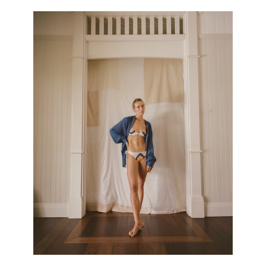 Young woman in a bikini and shirt stands in a doorway with natural light and a wood floor, exuding a casual yet confident vibe.
