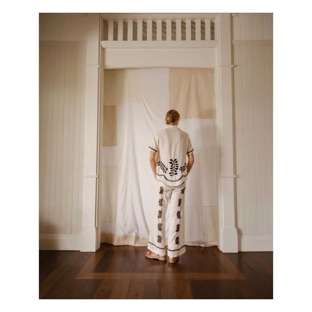 Back view of adult female in a cream-colored pant suit with dark trim, indoors in front of a draped cream fabric backdrop.