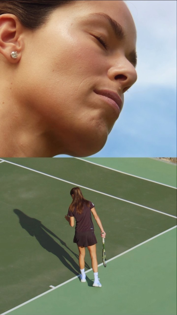 Female tennis player on a green outdoor court with a partial sky view in the background. Tennis player is seen from the back.