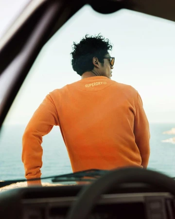 Man wearing an orange sweatshirt leaning against a car overlooking the ocean