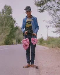 Young man wearing a baseball cap and blue jacket holding two pink flower garlands beside a rural road.