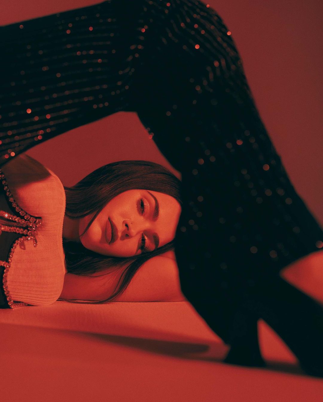 Portrait of a woman with long brown hair reclining in a studio with red lighting while someone in black sparkly pants stands over her.
