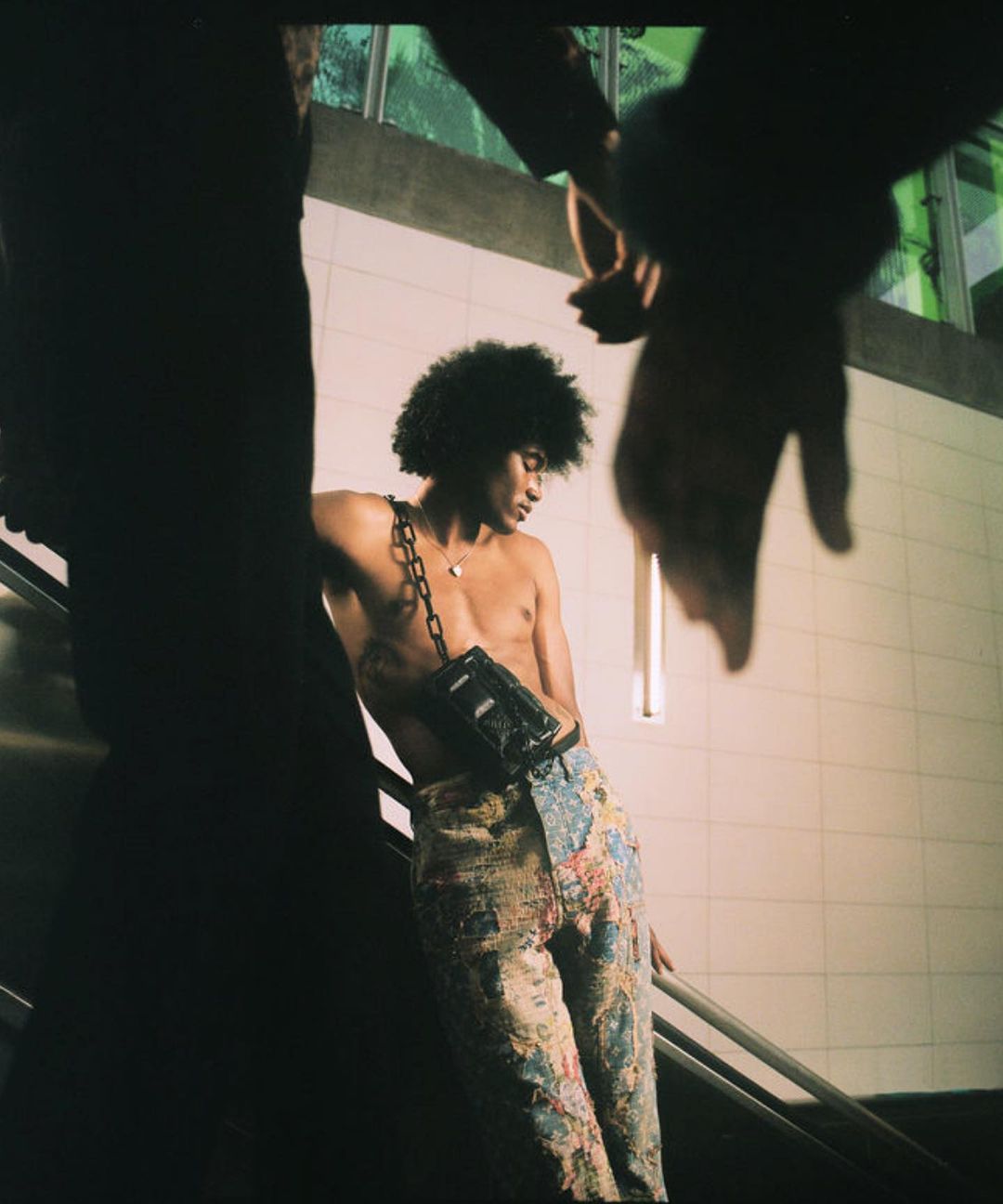 A shirtless African-American man with an afro and floral pants poses by an escalator in an urban setting with another person partially visible.