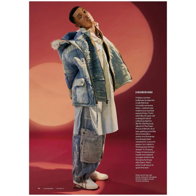 Jabari Banks poses in studio wearing a hooded denim jacket, white top, denim pants, and white sneakers against a red backdrop.