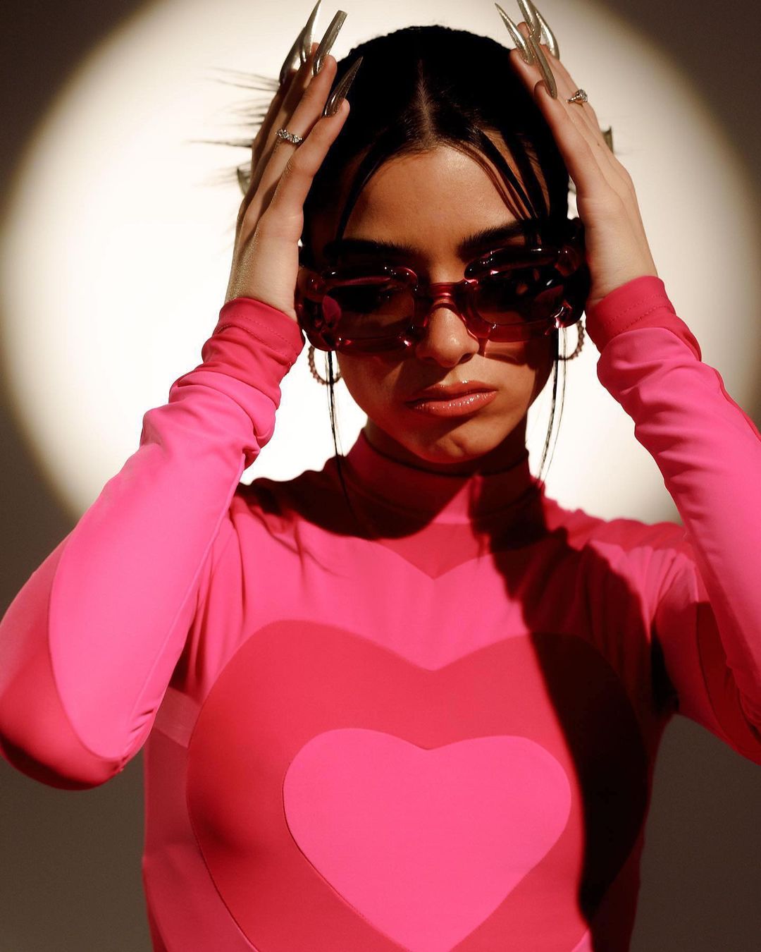 Stylish young woman with pink outfit, sunglasses, and long nails poses against a white backdrop with spotlight.