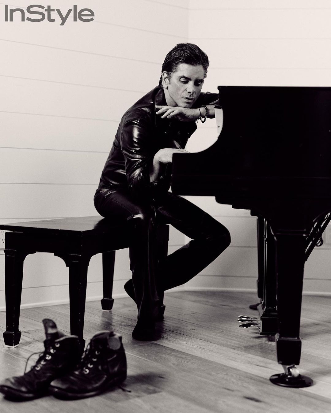 A man in a leather outfit is sitting and leaning on a black piano, with a pair of boots on the floor in a black and white photo.