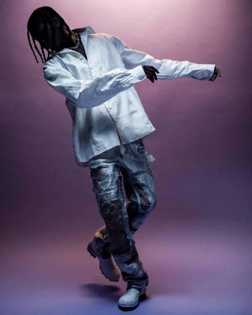 An adult male poses in an unbuttoned white shirt and textured silver pants on a light purple background, showcasing modern fashion.