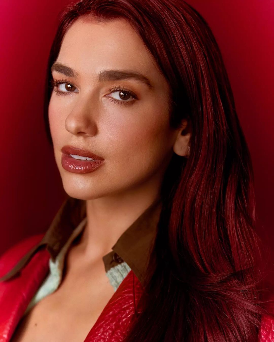 Close-up portrait of a brunette woman with burgundy hair, brown eyes, and glossy lips, wearing a red leather jacket against a red backdrop.