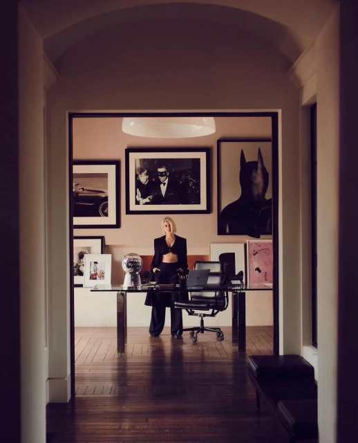 Adult female stands in an interior hallway with a glass desk and chair, artwork hanging on the wall in a home setting.