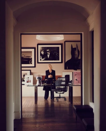 Adult female stands in an interior hallway with a glass desk and chair, artwork hanging on the wall in a home setting.