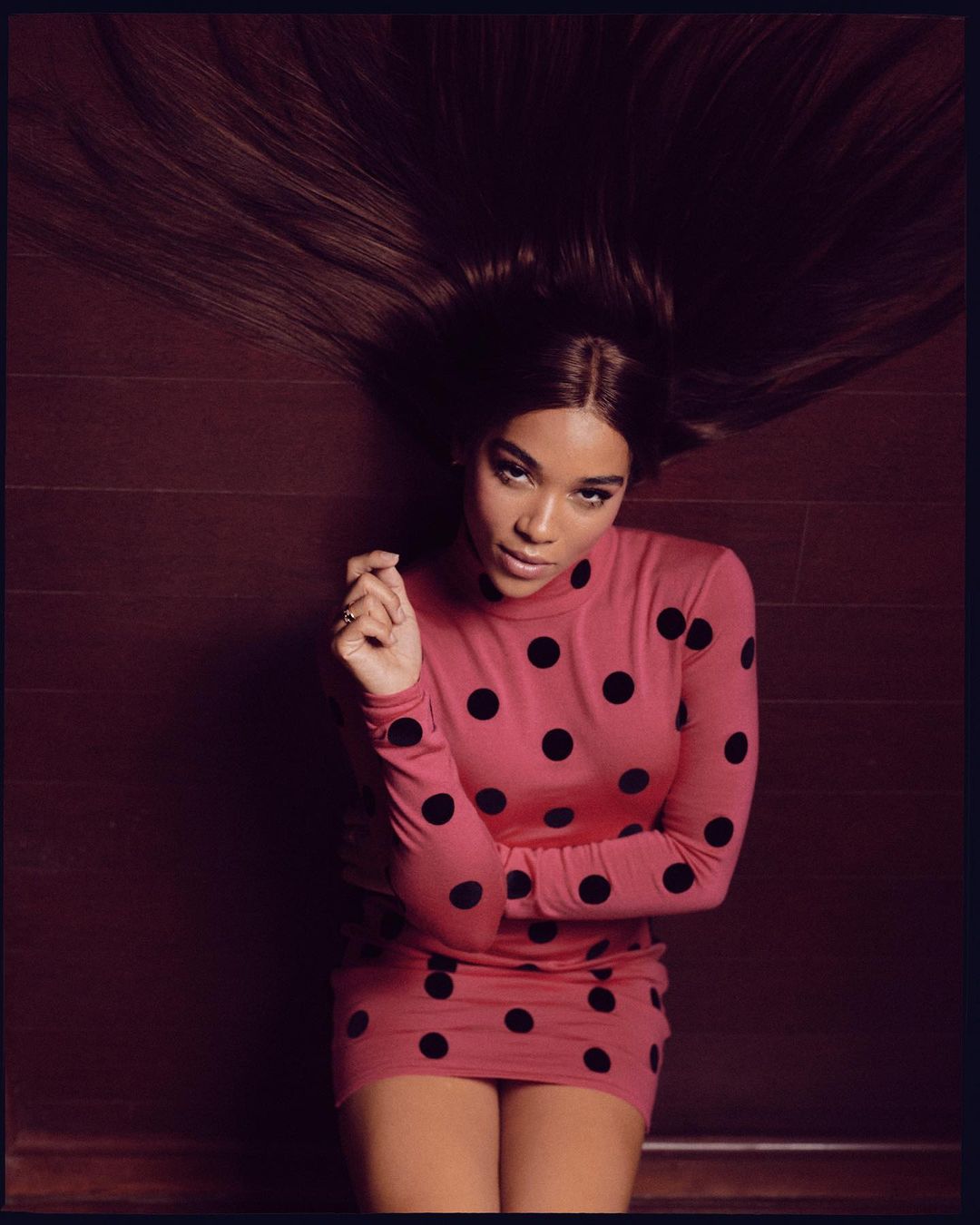Young woman in polka dot dress with long brown hair spread out on the floor, posing for fashion portrait in indoor setting.