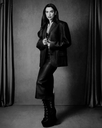 Monochrome portrait of Dua Lipa wearing a textured blazer, matching skirt, and platform boots in a studio setting.