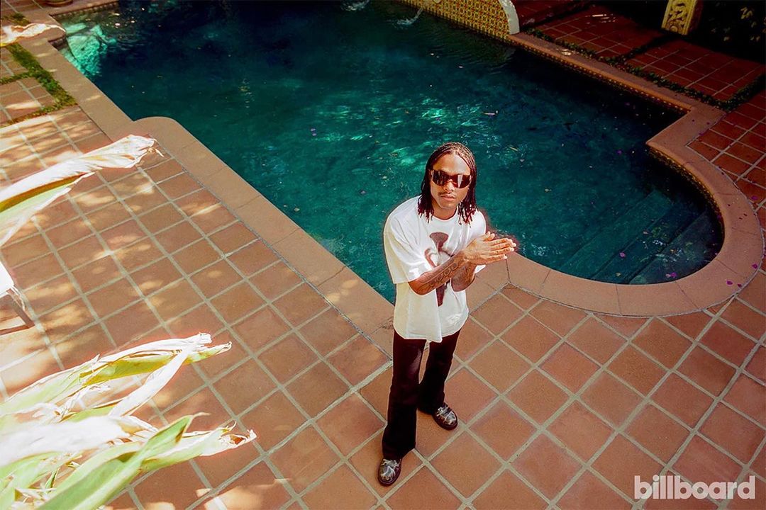 Man with dreadlocks stands beside a pool in sunglasses, wearing a white t-shirt and black pants, with the Billboard logo at the bottom.