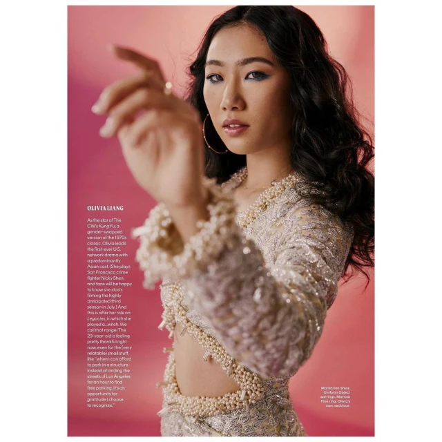 Portrait of an Asian woman wearing a beaded and sequined dress with cutouts, hoop earrings, and ring, against a pink background.
