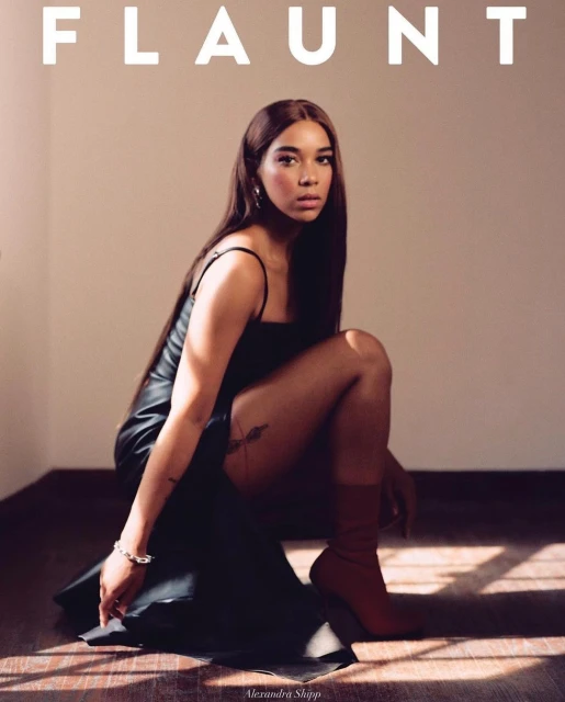 Alexandra Shipp poses in a black dress and brown boots for Flaunt magazine. The portrait is captured indoors with soft, diffused lighting.