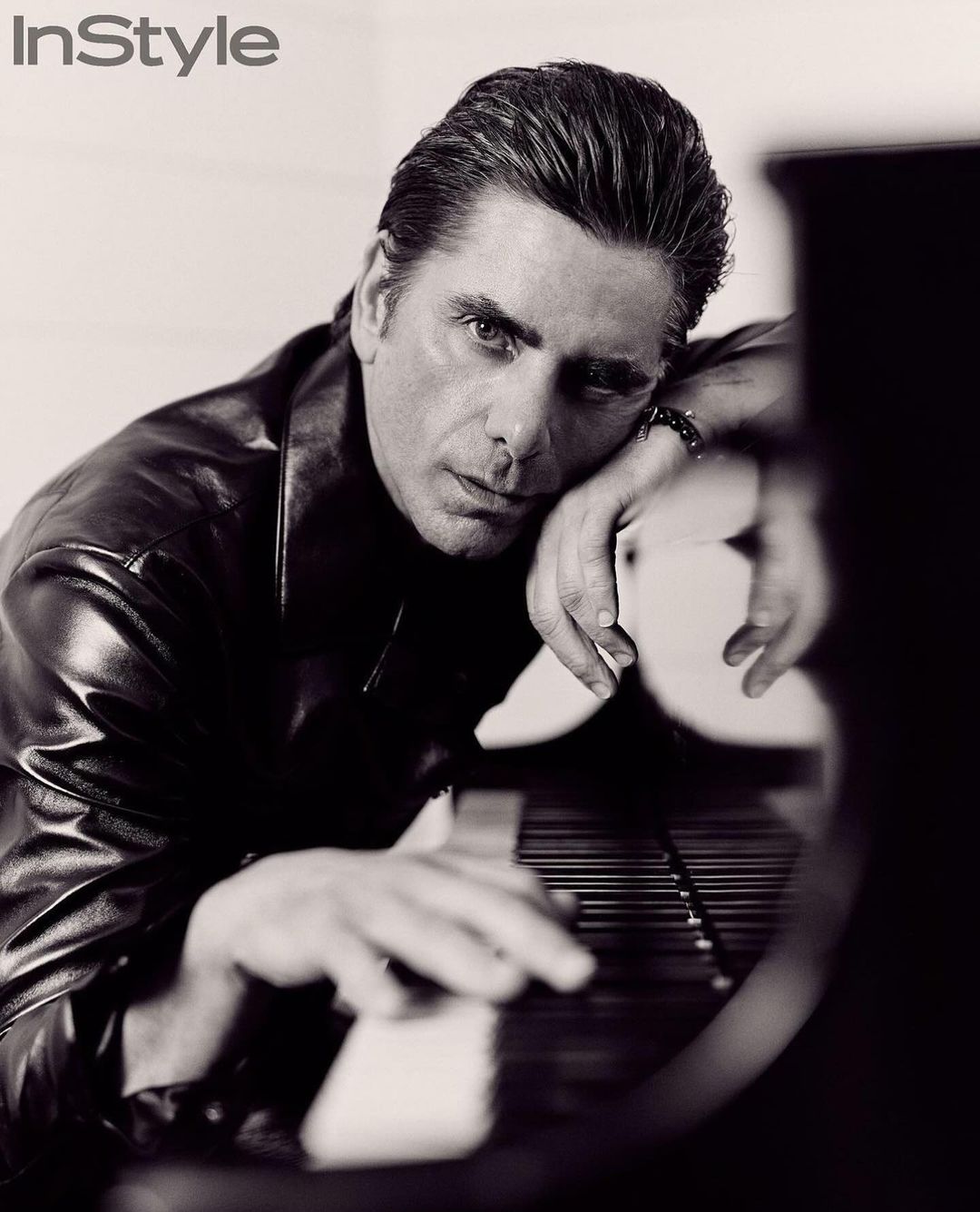 Black and white portrait of a man with dark hair and a leather jacket, resting his hands on a piano keyboard, with InStyle logo.