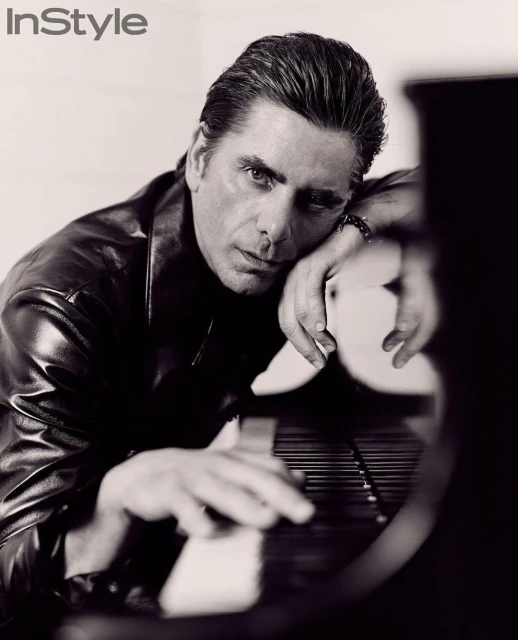 Black and white portrait of a man with dark hair and a leather jacket, resting his hands on a piano keyboard, with InStyle logo.