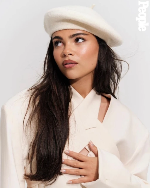 Portrait of a smiling young woman wearing a white beret and cream-colored jacket in a photo studio with a white background.