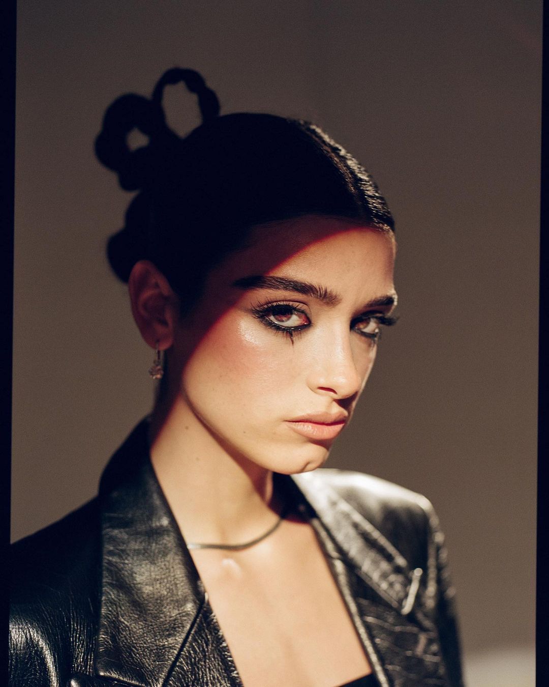 Portrait of a young adult woman with dark hair and heavy eye makeup, wearing a black leather jacket against a neutral backdrop.