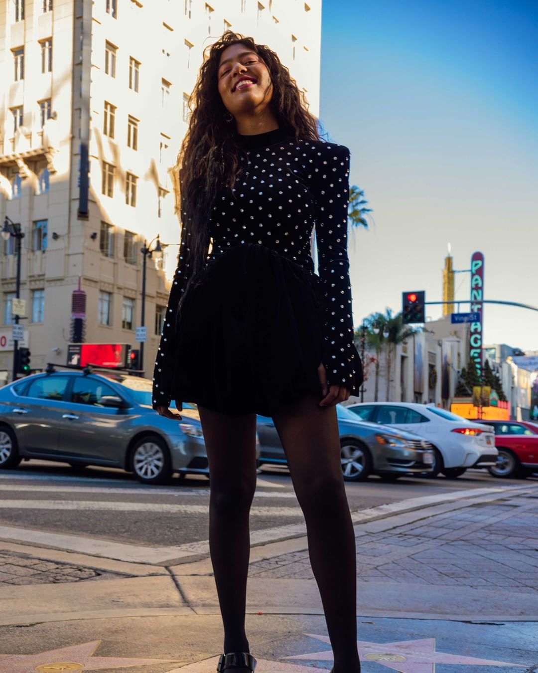 Smiling woman in long-sleeved polka dot top and black skirt poses on a crosswalk with city buildings and traffic in the background.