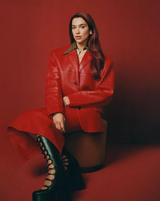 A redhead woman poses in a studio wearing a red leather suit and high platform boots. The photograph is set against a monochrome red backdrop.