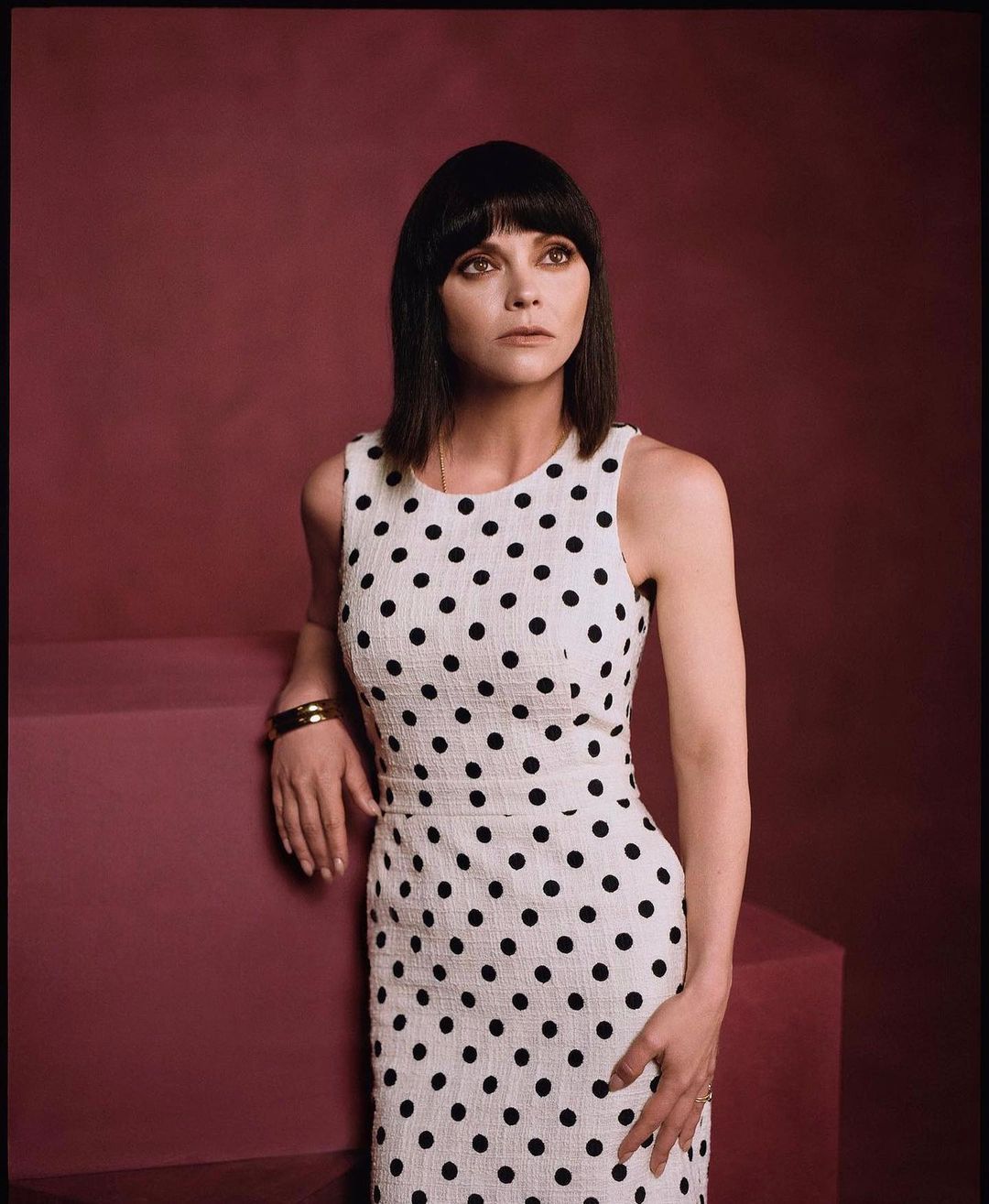 Christina Ricci poses in a white polka dot dress against a mauve backdrop, wrist adorned with a gold cuff bracelet.