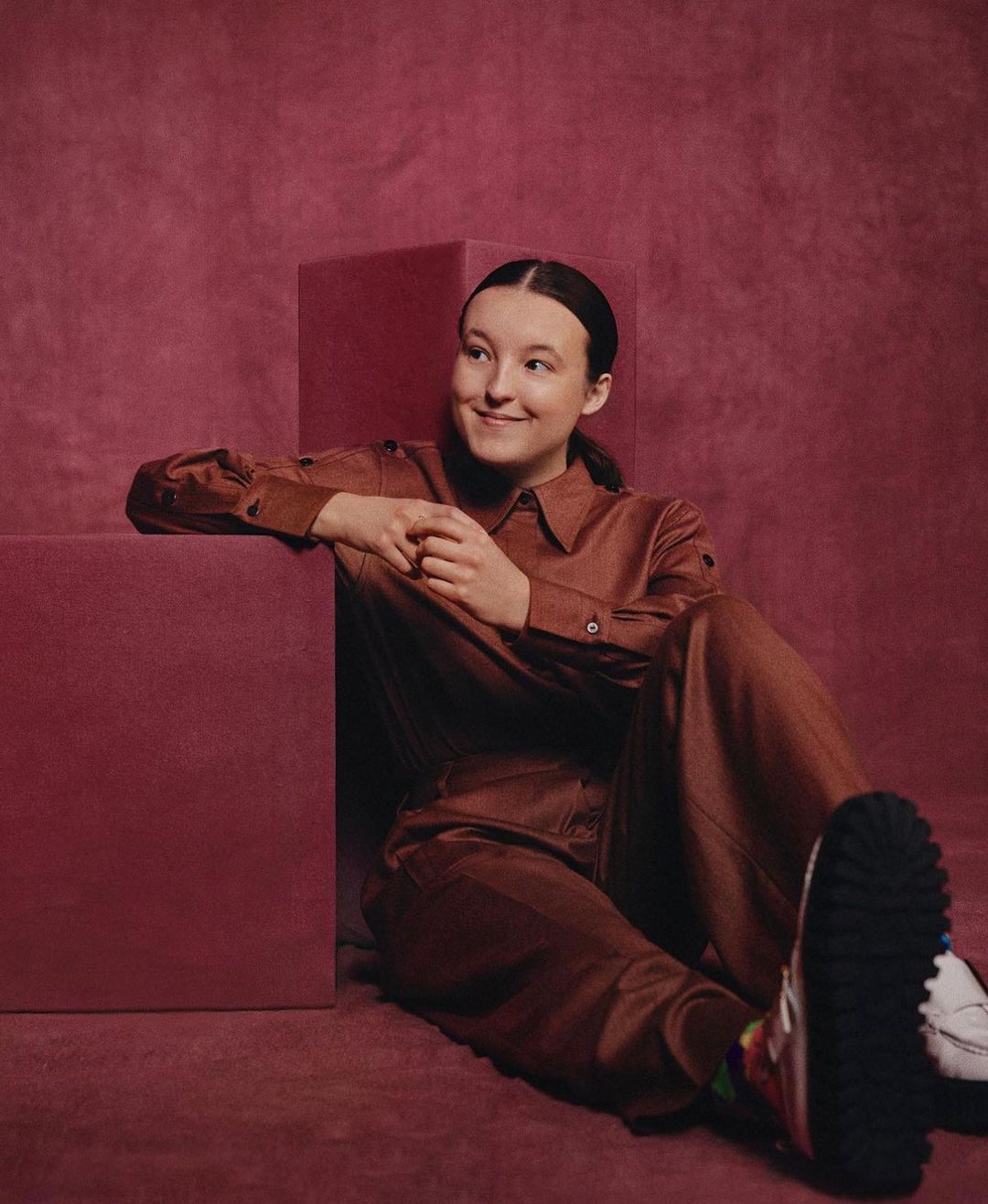 Portrait of Bella Ramsey wearing a brown jumpsuit leaning against a brown cube in a photo studio with a matching brown backdrop.