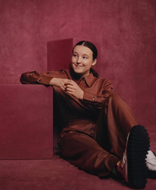 Portrait of Bella Ramsey wearing a brown jumpsuit leaning against a brown cube in a photo studio with a matching brown backdrop.