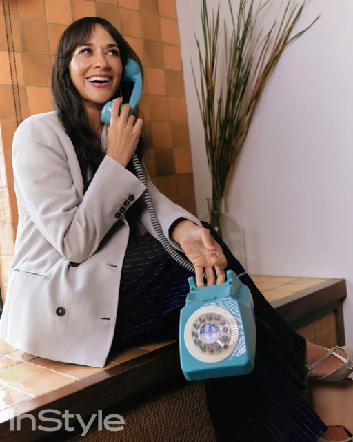 A woman in a blazer sits holding a light blue rotary phone in a casual indoor setting with tan tile and decorative grass.