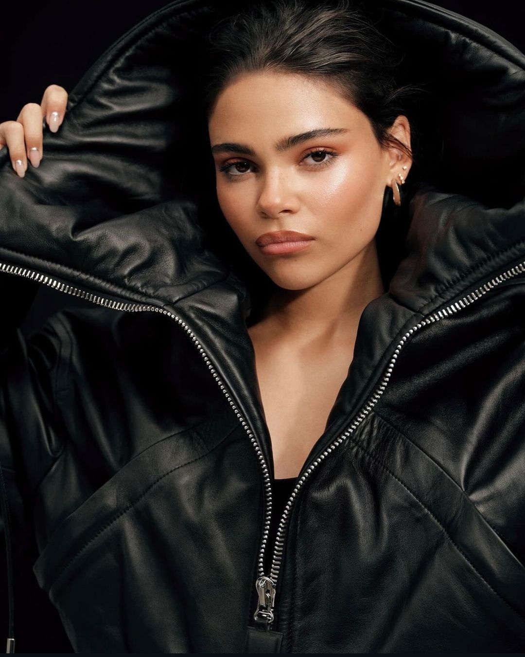 Close-up studio portrait of a young adult woman with light skin wearing a black leather jacket with a large hood, looking at camera.