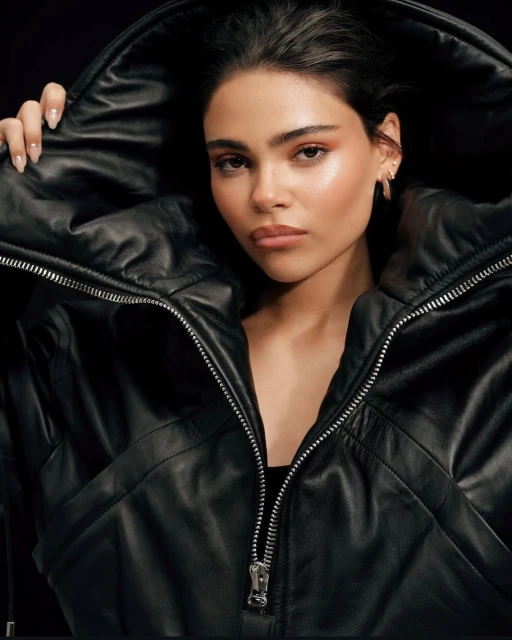 Close-up studio portrait of a young adult woman with light skin wearing a black leather jacket with a large hood, looking at camera.