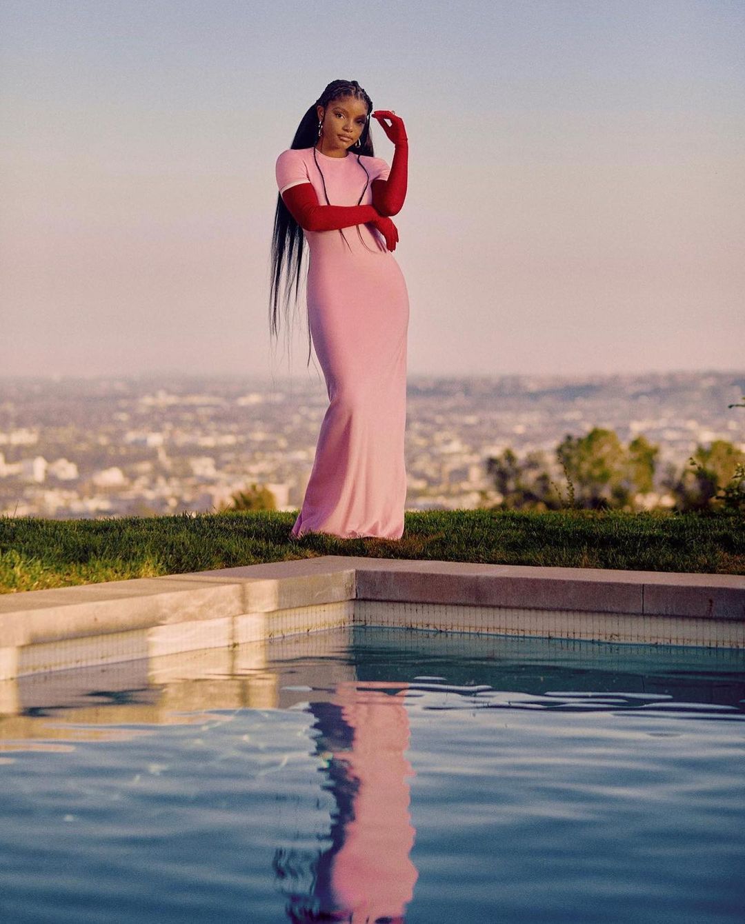 A woman in a pink dress and red gloves poses near a pool with a city and light pink sky in the background at golden hour.