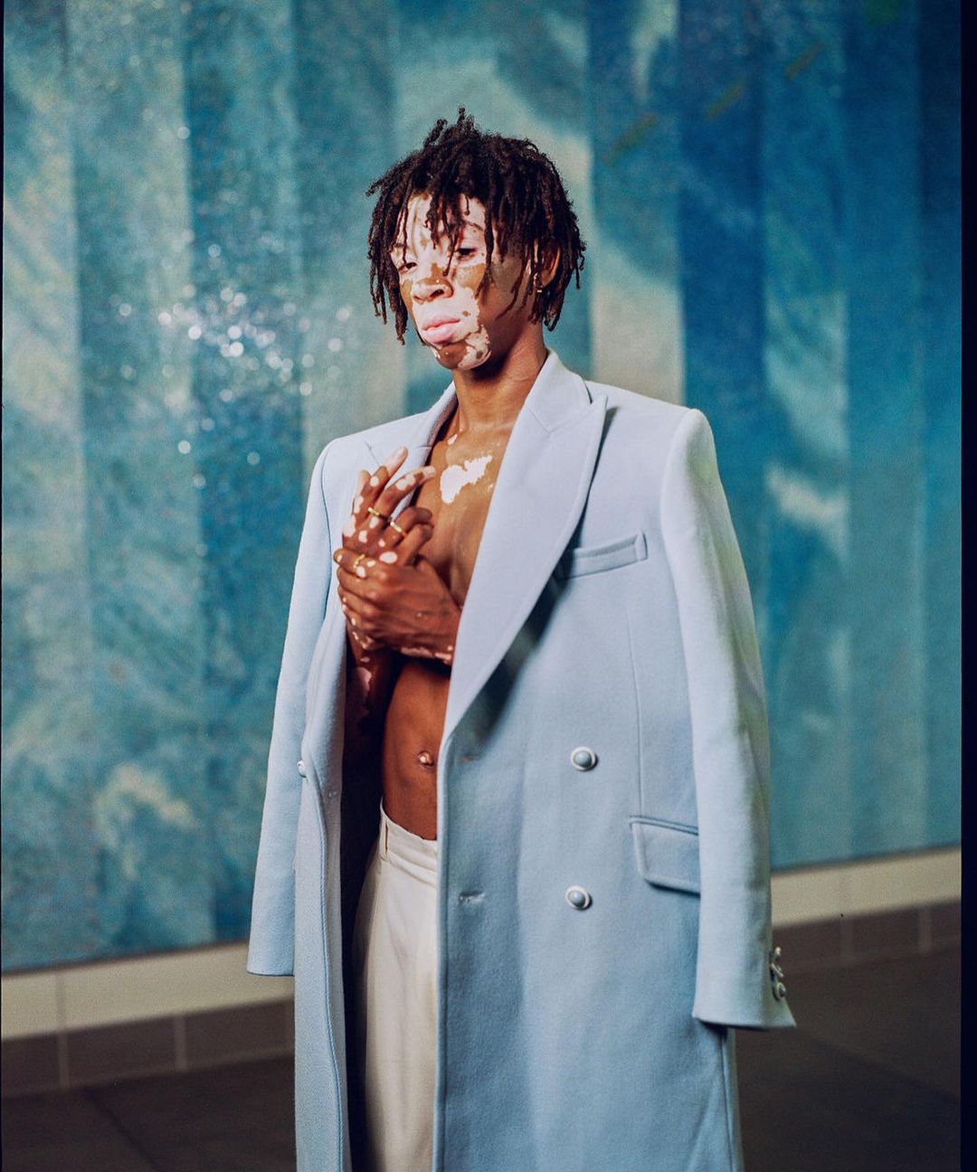 Portrait by Alex Harper of a man wearing an open light blue double-breasted coat against a blue tiled wall in Los Angeles.