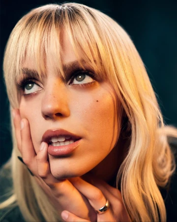 Close-up portrait of a blonde woman with bangs looking upward, resting her chin on her hands with fingers near her lips.