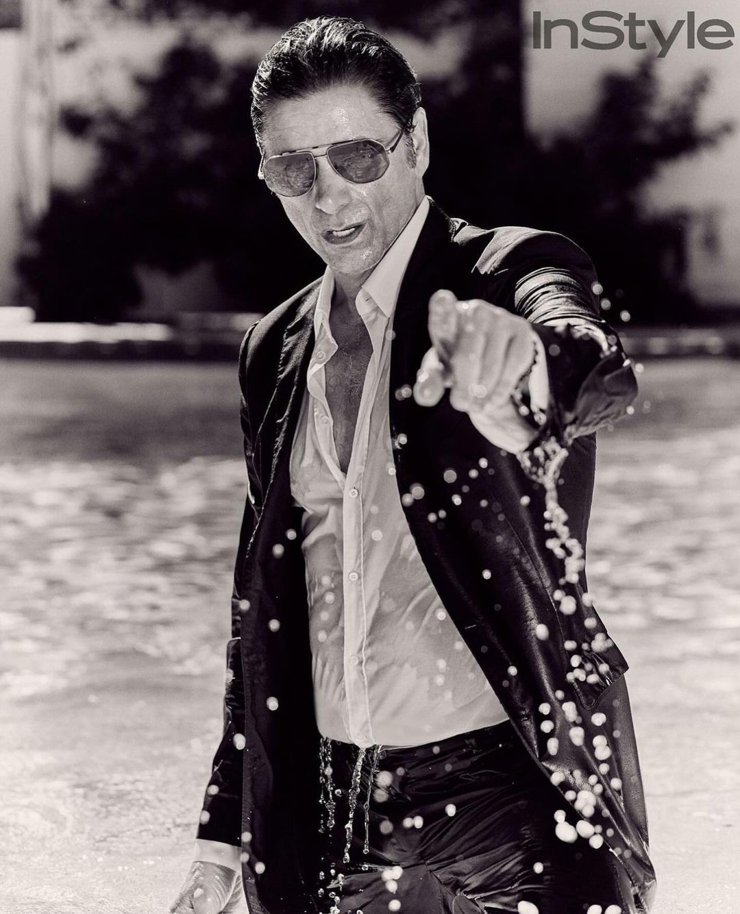 Monochrome portrait of a wet adult male in a suit and sunglasses pointing at the camera in an outdoor water setting.