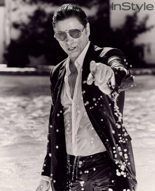 Monochrome portrait of a wet adult male in a suit and sunglasses pointing at the camera in an outdoor water setting.