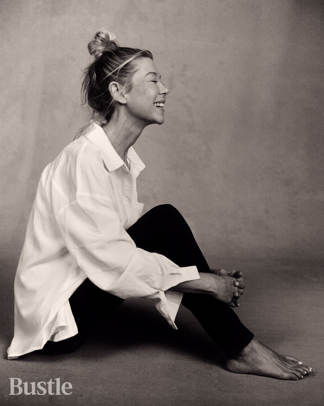 Monochrome portrait of a smiling woman with blonde hair, wearing a white button-down shirt and black pants, sitting on the floor.