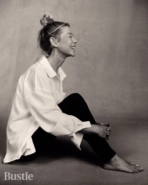 Monochrome portrait of a smiling woman with blonde hair, wearing a white button-down shirt and black pants, sitting on the floor.