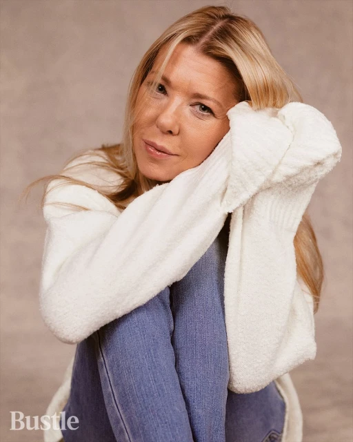 Portrait of a blonde woman in a white sweater and blue jeans sitting indoors, looking at the camera with a slight smile, with soft lighting.