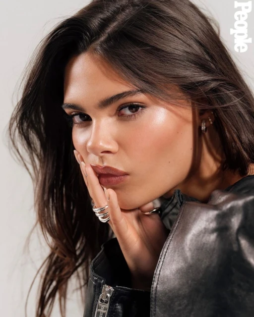 Close-up portrait of a brunette woman wearing a black leather jacket and silver rings posing in front of a neutral background.