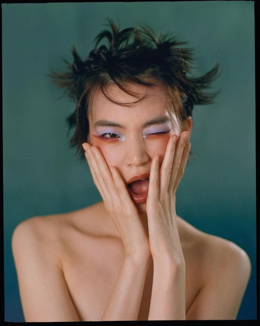 Close-up studio portrait of a young adult woman with wild hair and creative makeup, posing with hands on her cheeks, against teal backdrop.