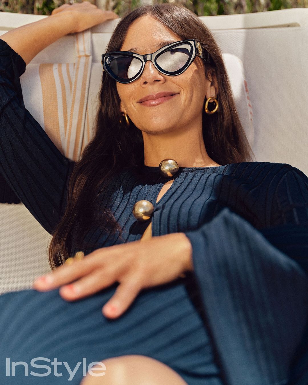 Brunette woman wearing sunglasses and a blue, button-up dress while reclining on a striped chaise lounge outdoors in natural light.